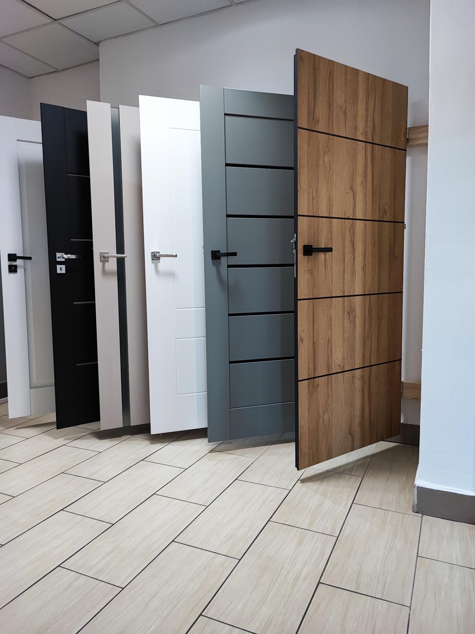 Ekspozycja modeli drzwi wewnętrznych w różnych kolorach: biały, czarny, szary i drewnopodobny z czarnymi klamkami, ustawione w rzędzie na jasnej podłodze.