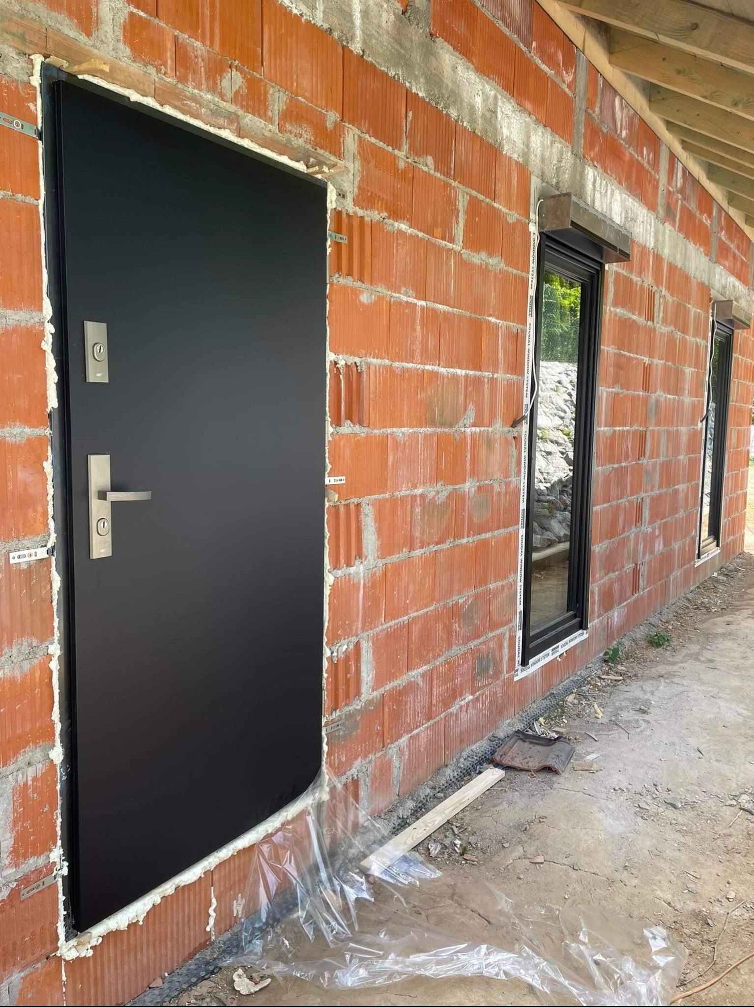 Montaż czarnych drzwi zewnętrznych i okien z roletami w surowym stanie budynku z czerwonej cegły, widoczne pianka montażowa i folia ochronna.