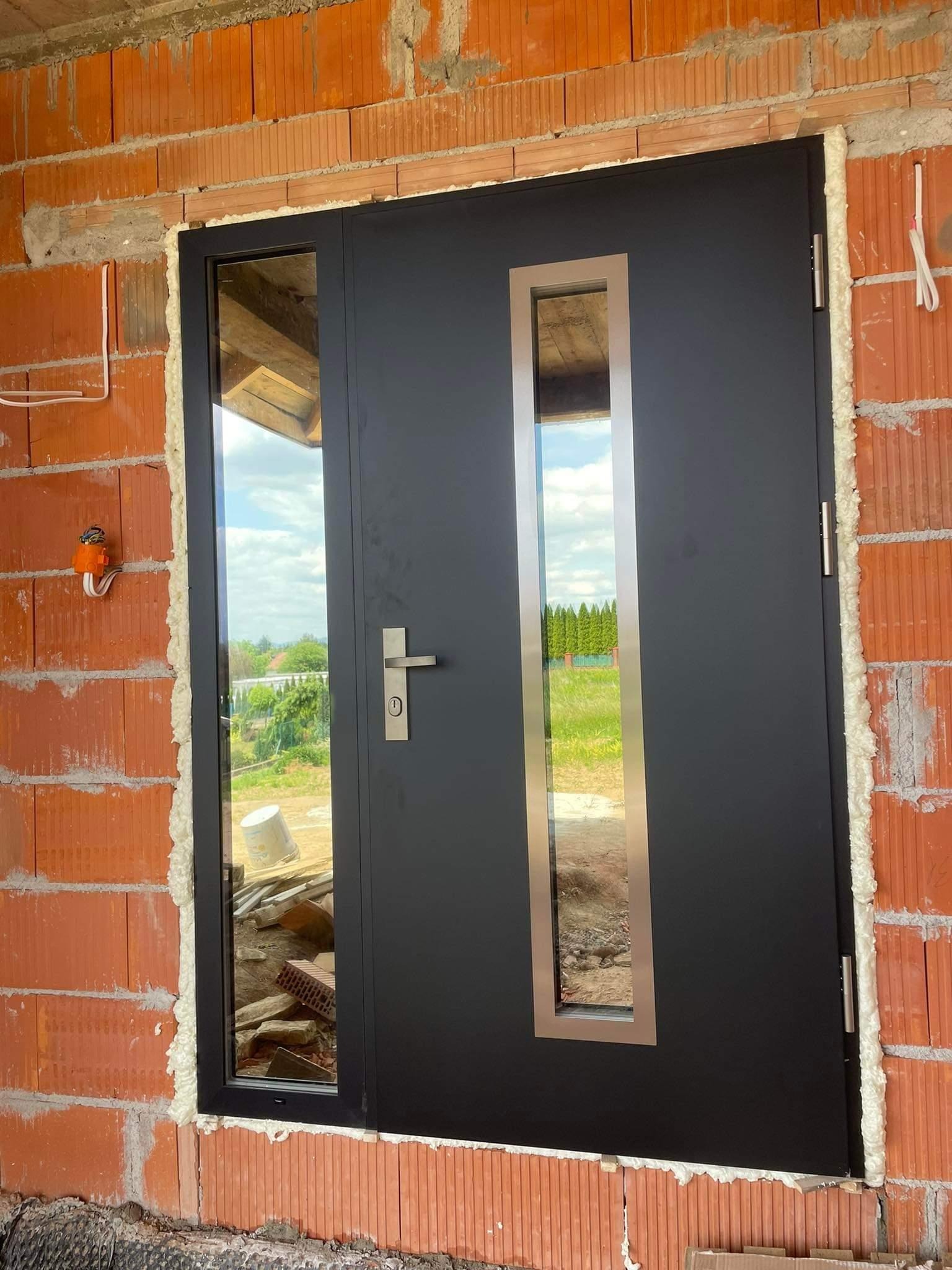 Ciemne drzwi zewnętrzne z przeszkleniami, zamontowane w surowym murze z czerwonej cegły, uszczelnione pianką montażową, widok na zewnątrz przez szyby.
