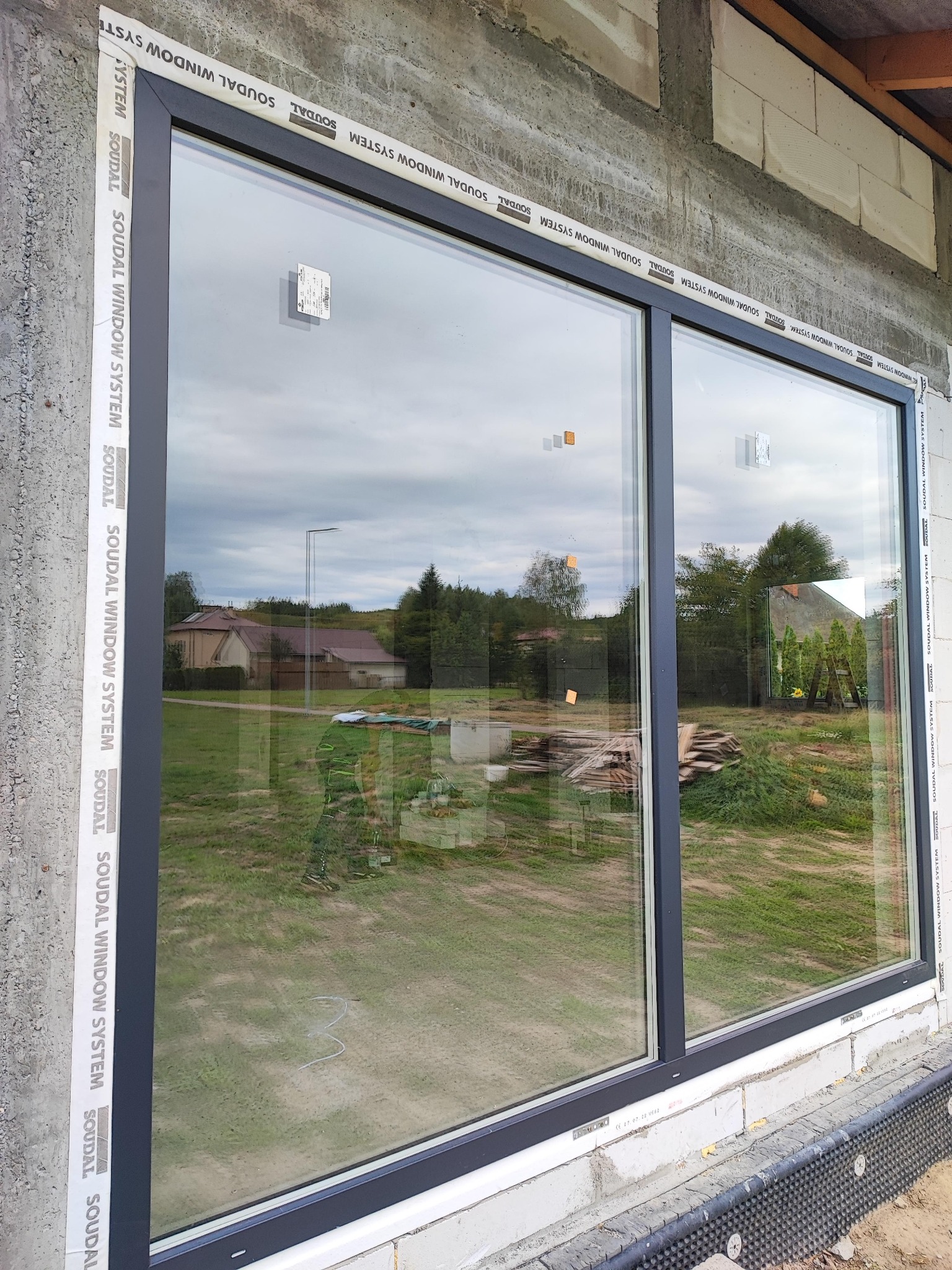 Duże, dwuczęściowe okno z szarą ramą zamontowane w surowej ścianie budynku w trakcie budowy, z widoczną izolacją i taśmami uszczelniającymi Soudal Window System.