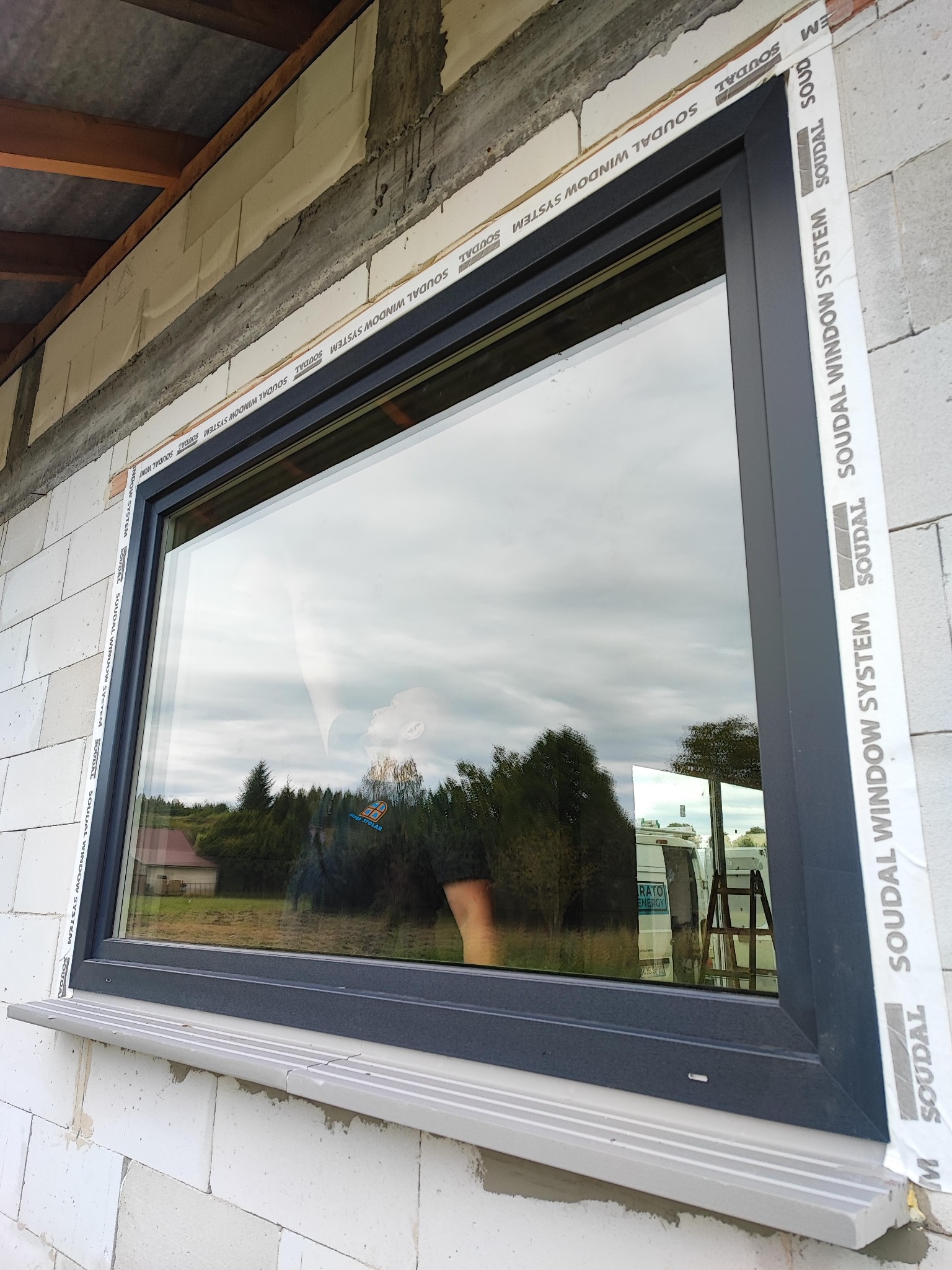 Nowo zamontowane okno PCV w kolorze antracytowym, widoczne białe bloczki konstrukcyjne i taśma uszczelniająca Soudal Window System, z odbiciem krajobrazu i osoby w szybie.