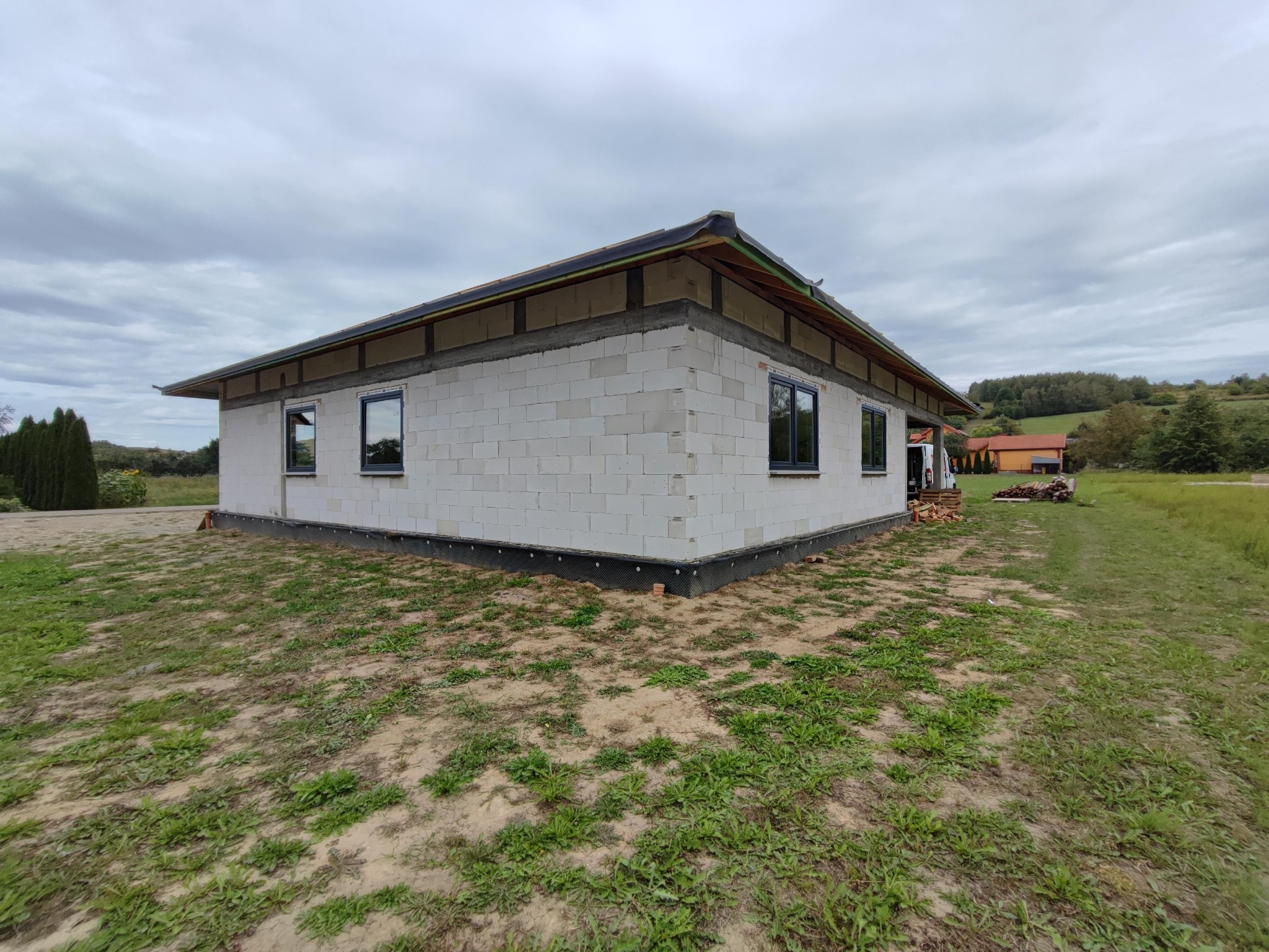 Budynek w trakcie budowy z zainstalowanymi oknami, widoczne ściany z bloczków betonowych, zabezpieczenie fundamentów i konstrukcja dachu, na zewnątrz trawnik i krajobraz wiejski.