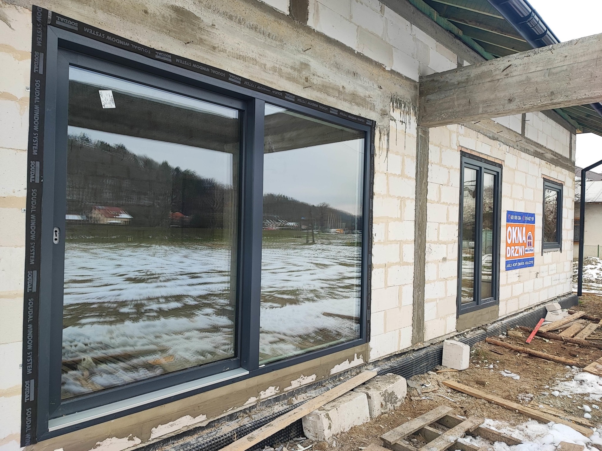 Duże okno przesuwne z szarą ramą w surowym stanie budynku, widoczna izolacja i teren z resztkami śniegu, na ścianie baner reklamowy okien i drzwi.