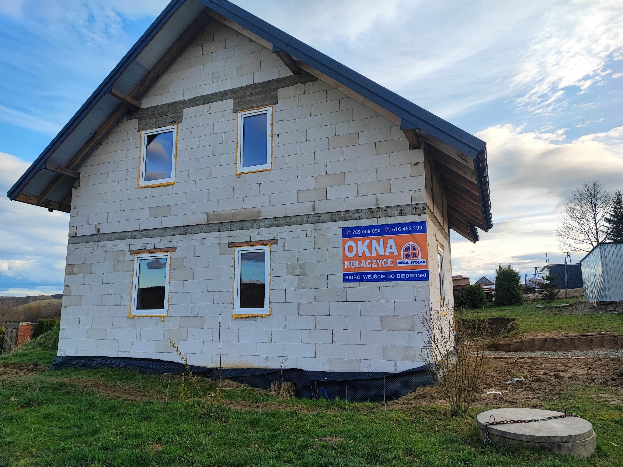 Surowy budynek z białego bloczka z zamontowanymi oknami PCV, widoczna pianka montażowa wokół okien, na ścianie reklama firmy 'Okna Kołaczyce', teren wokół budynku w trakcie prac ziemnych.