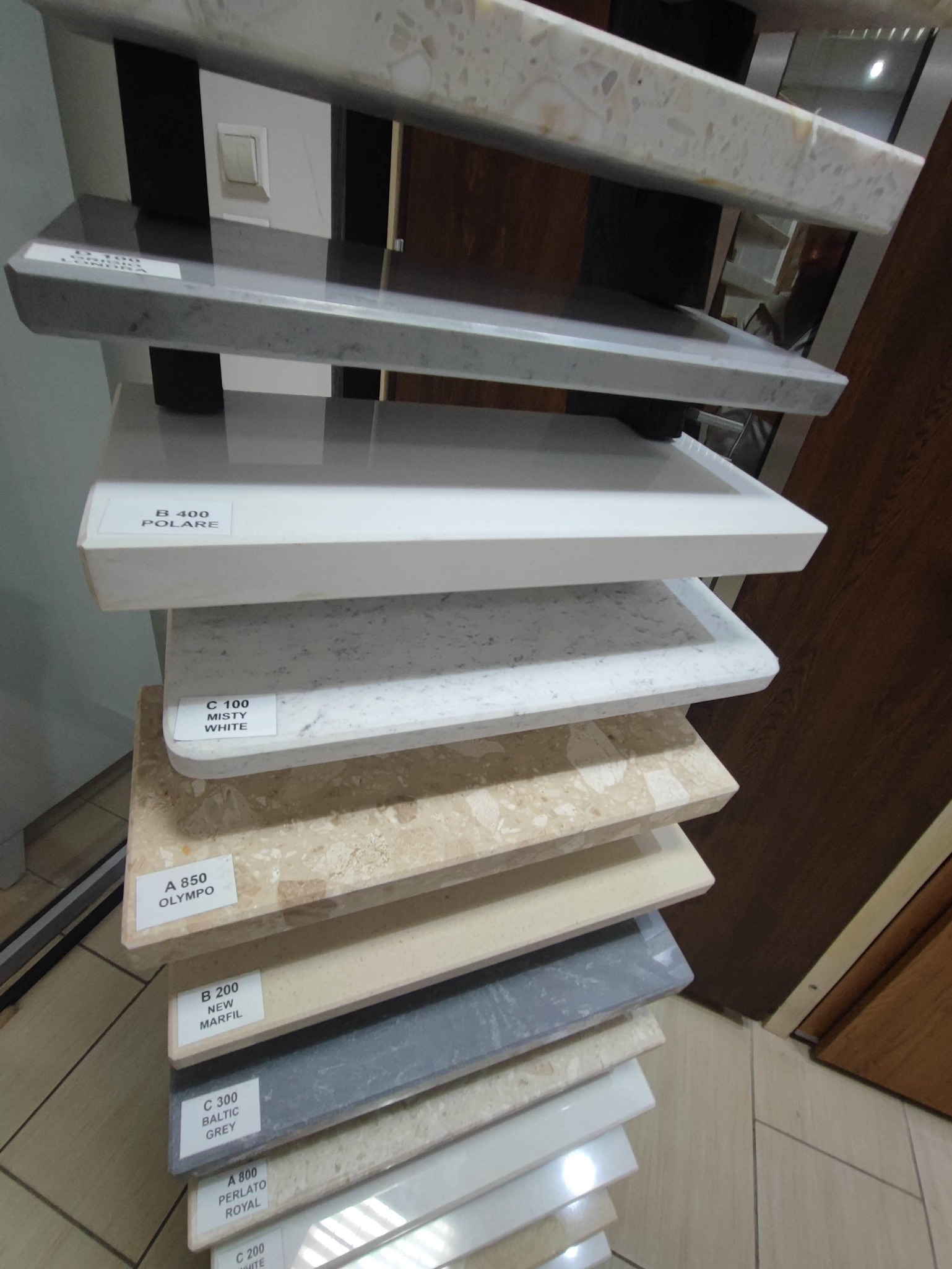 Ekspozycja próbek różnych kolorów i wzorów parapetów, ułożonych jeden na drugim, z widocznymi etykietami z nazwami: Olympo, New Marfil, Baltic Grey, Perlato Royal, Misty White, Polare.