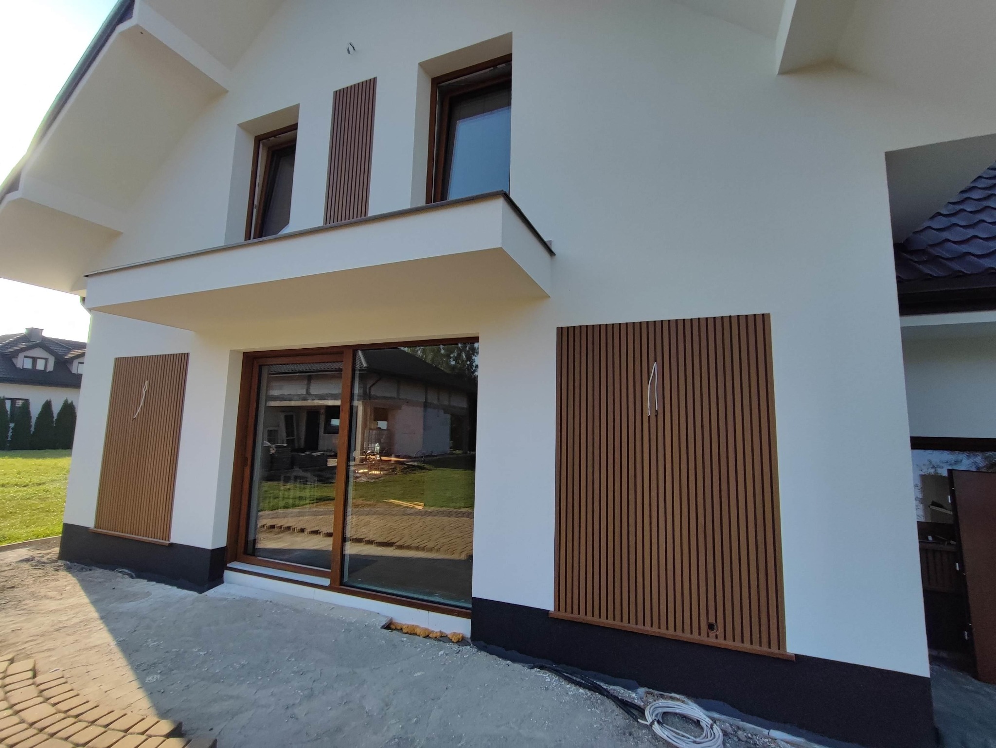 Nowoczesny dom z oknami PCV w brązowych ramach i dekoracyjnymi panelami ściennymi z pionowych listew w kolorze drewna, widok z zewnątrz na plac budowy z przewodami.