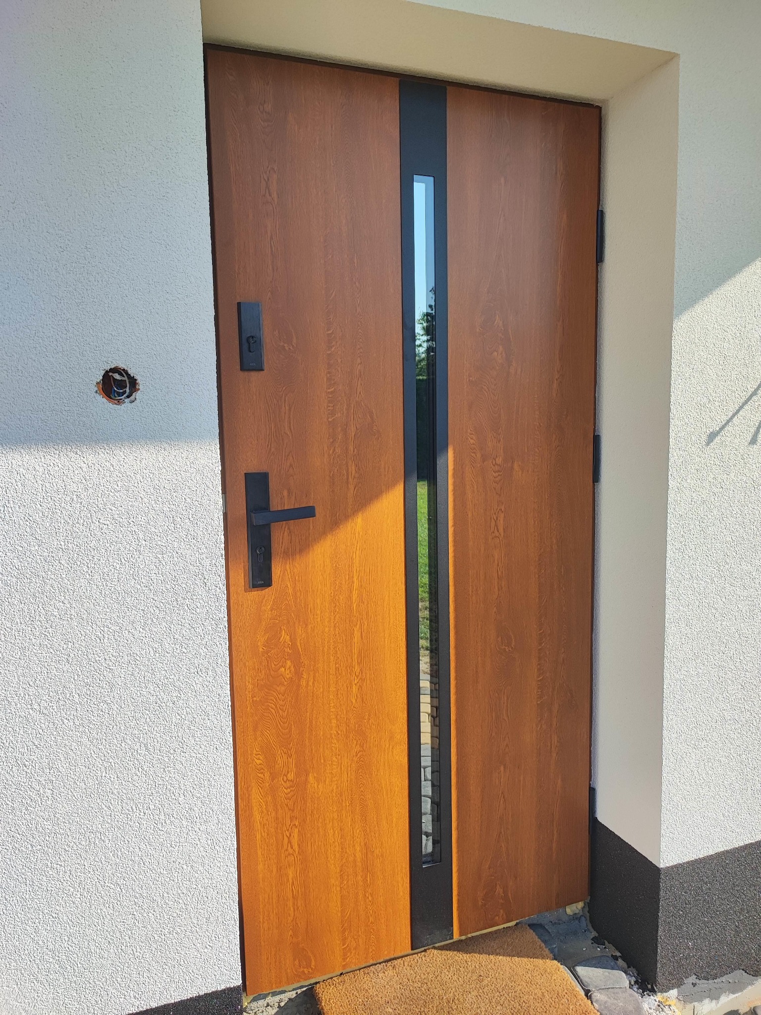 Nowoczesne drzwi zewnętrzne z drewnopodobnym wykończeniem i czarnymi elementami, w tym długi uchwyt i wąskie przeszklenie, zamontowane w jasnym, otynkowanym murze z widocznym otworem na instalację.