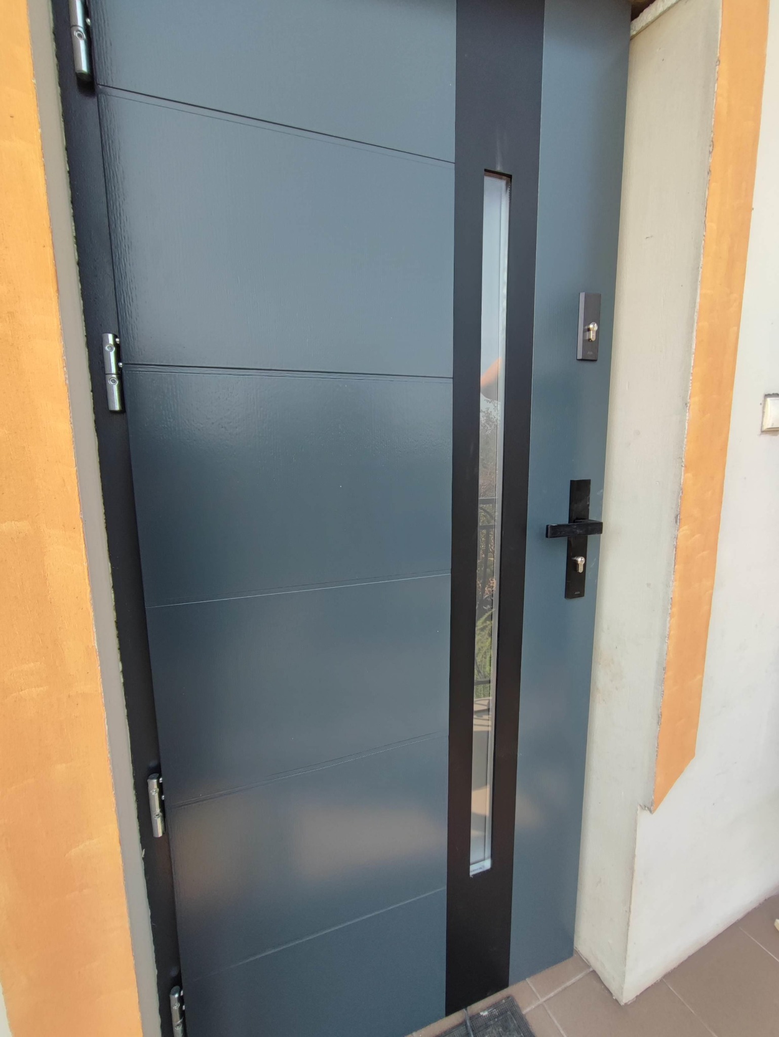 Szare drzwi wejściowe z poziomymi panelami, czarną wstawką z szybą i klamką, zamontowane w ościeżnicy przy pomarańczowym narożniku ściany.