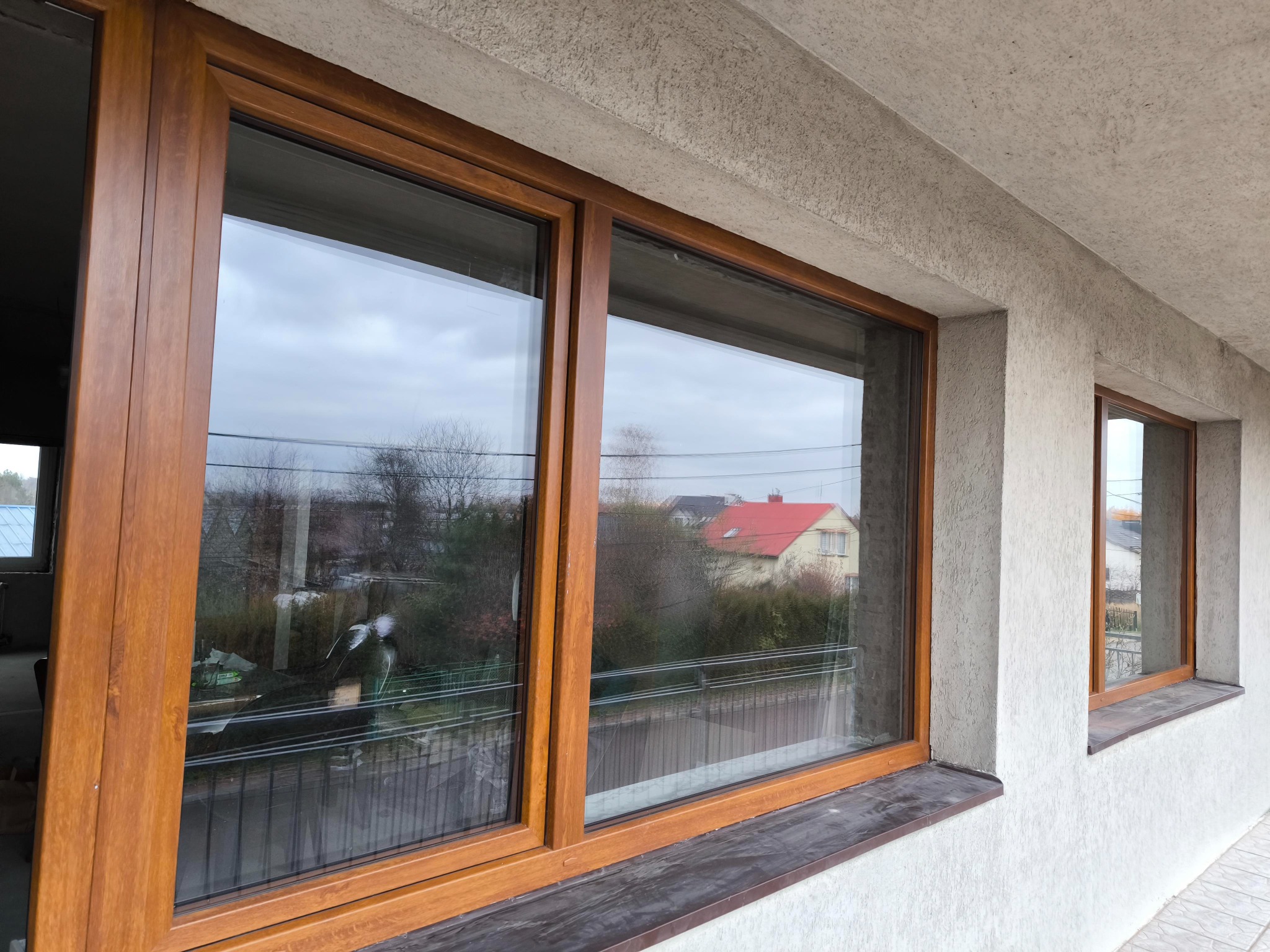 Dwa okna z brązowymi ramami zamontowane w elewacji budynku o strukturze baranka, widok na ogród i dom z czerwonym dachem.