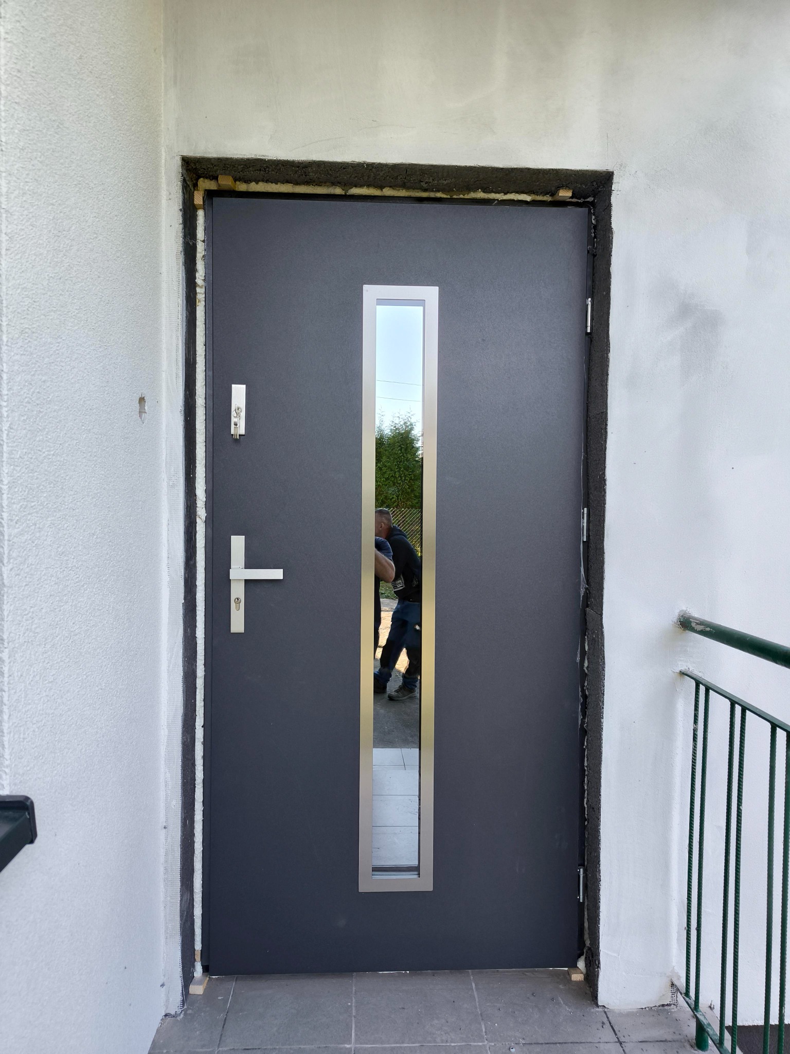 Montaż szarych drzwi wejściowych z podłużnym przeszkleniem, widoczne pianka montażowa i elementy uszczelniające wokół ościeżnicy, odbicie montera w szybie.