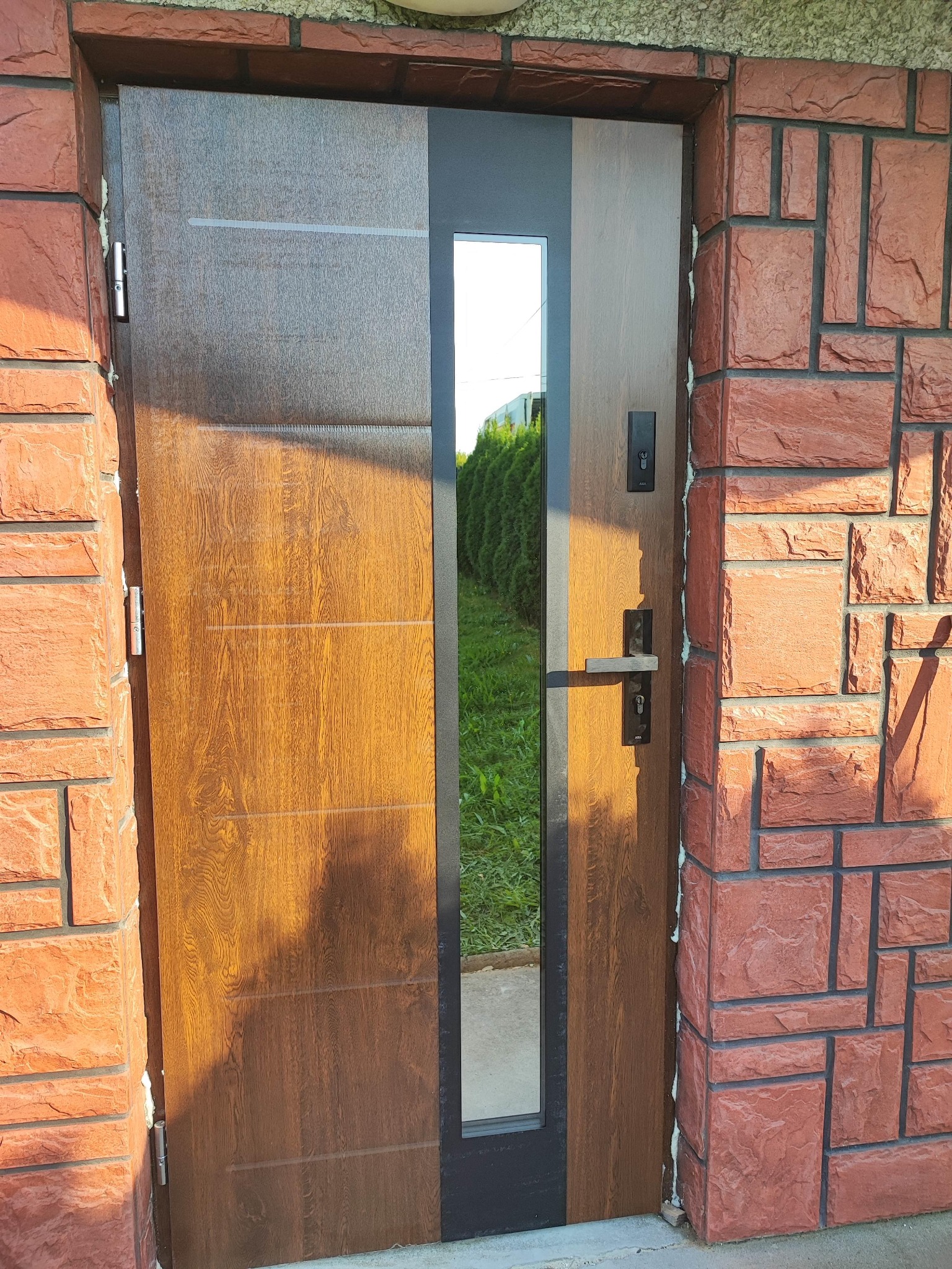 Brązowe drzwi wejściowe z szybą, zamontowane w czerwonej ceglanej elewacji, widoczne ślady pianki montażowej.