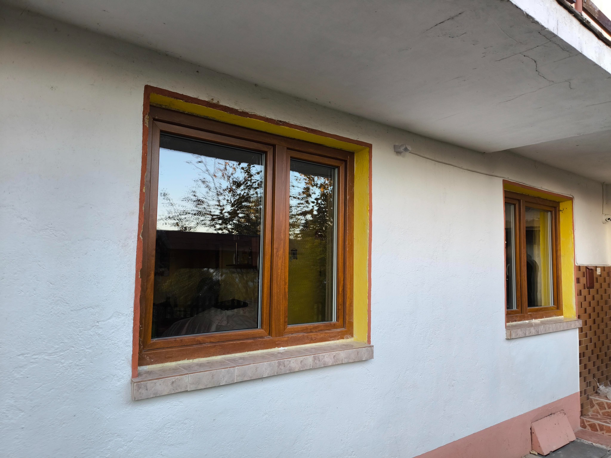 Dwa brązowe okna z ramami w odcieniach żółci i pomarańczy, osadzone w białej, teksturowanej ścianie zewnętrznej budynku, z widocznym odbiciem drzew w szybach.