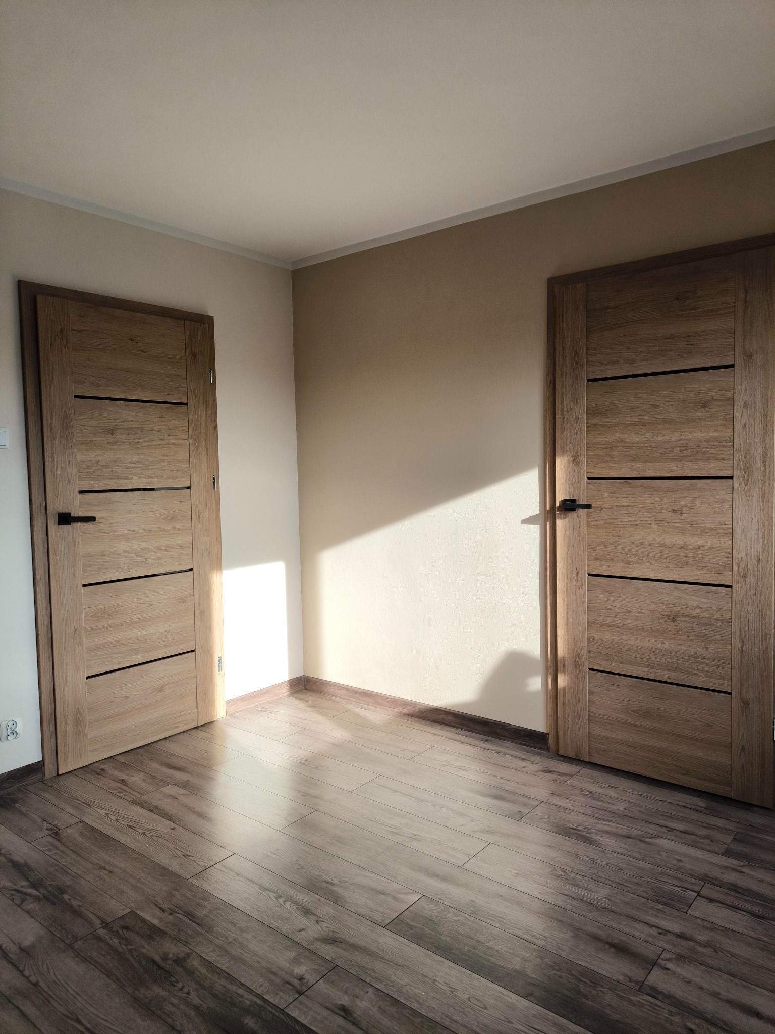 Dwa drewniane drzwi wewnętrzne z czarnymi klamkami, rozmieszczone po przeciwnych stronach pokoju z panelami podłogowymi, oświetlone padającym światłem słonecznym.