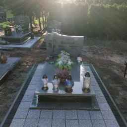 Granitowa oprawa grobu z szarych płyt i ciemnych obrzeży, z zapalonymi zniczami i kwiatami, oświetlona promieniami słonecznymi.