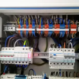 Otwarta skrzynka elektryczna z licznymi przewodami, bezpiecznikami i złączkami, widoczne oznaczenia na elementach, wewnątrz biała obudowa.