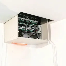 Szafka sterująca mini instalacją SMART MIESZKANIA, zamontowana pod sufitem, z widocznymi przewodami i elementami elektroniki wewnątrz, oświetlenie LED i białe ściany w tle.