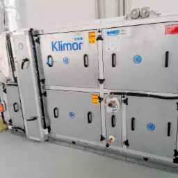 Centrala wentylacyjna Klimor w pomieszczeniu technicznym, widoczne przewody wentylacyjne w izolacji, elementy instalacji wodnej i elektrycznej.