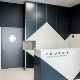 Nowoczesna recepcja w kolorach biało-granatowych z logo 'TruFey Watch Service', podświetlana od dołu, z drzwiami z okrągłym okienkiem po lewej stronie i wzorem geometrycznym na ścianie.