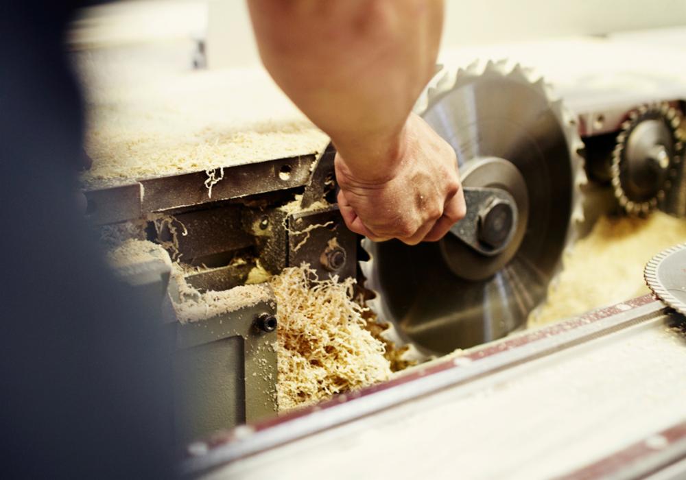 Ręka pracownika obsługującego piłę tarczową do cięcia drewna, z widocznymi wiórami i pyłem drzewnym.
