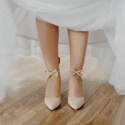 Panna młoda w kremowych szpilkach z kokardkami, widoczne spod białej sukni na parkiecie.