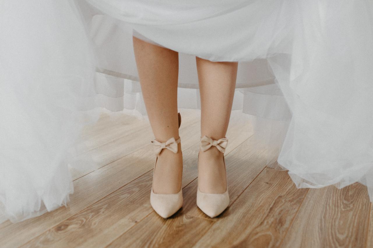 Panna młoda w kremowych szpilkach z kokardkami, widoczne spod białej sukni na parkiecie.