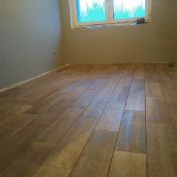 Ułożona podłoga z płytek imitujących drewno w nowym pokoju z szarą ścianą i oknem, widoczny poziomica na parapecie.