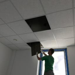 Armar - Pracownik w Nowym Dworze Gdańskim montuje kaseton sufitowy w pomieszczeniu z oknem, widoczny fragment ściany i biała kratownica sufitu podwieszanego.