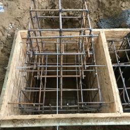 Armar - Zbrojenie fundamentu w drewnianym szalunku, widoczne pręty zbrojeniowe połączone drutem, przygotowane do zalania betonem. Częściowo zalany beton w sąsiedniej sekcji szalunku.