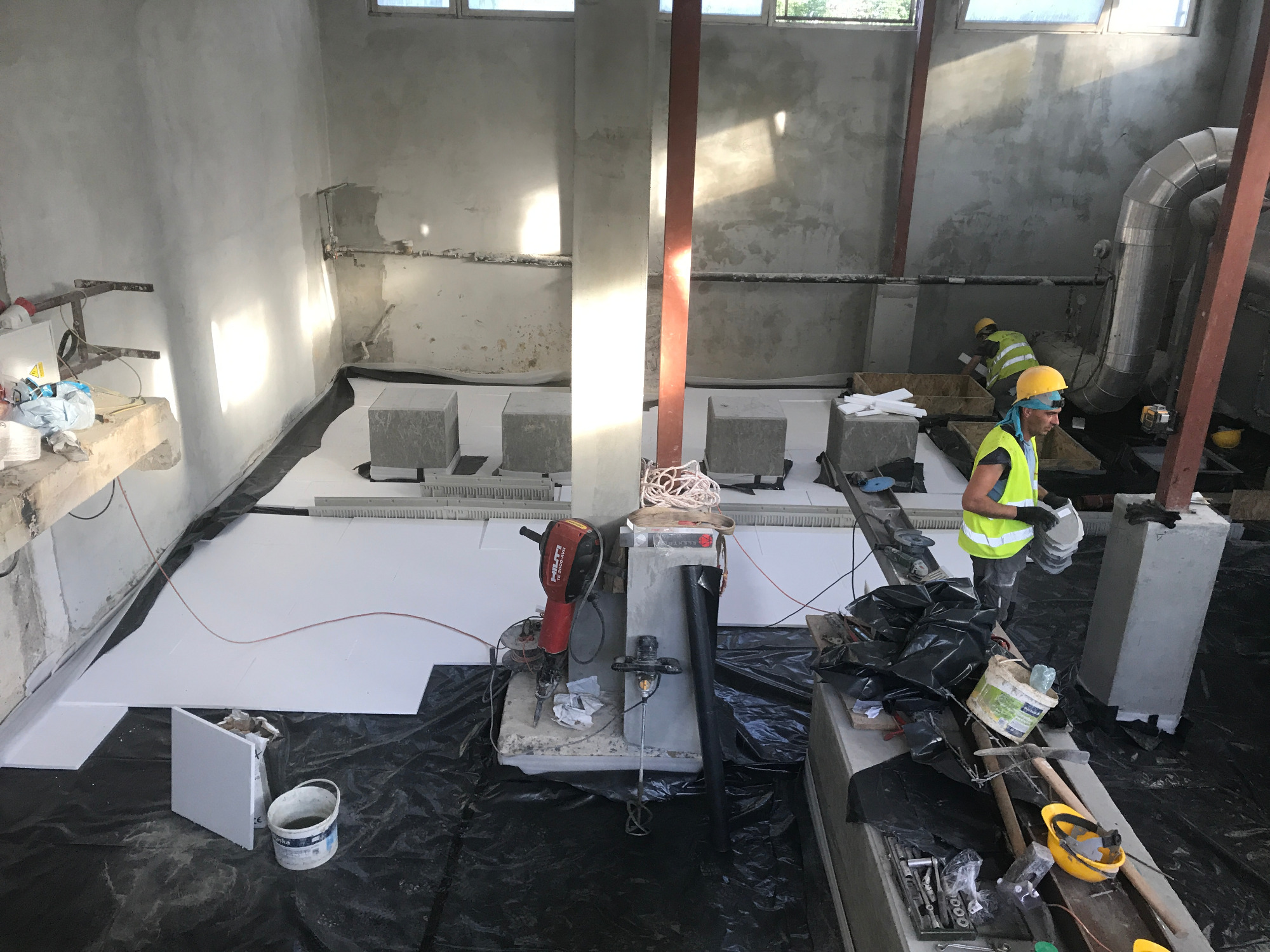 Wnętrze remontowanej stacji podnoszenia ciśnień: pracownicy układają izolację z białych płyt styropianowych na czarnej folii, widoczne narzędzia, betonowe podpory i fragment instalacji wentylacyjnej.