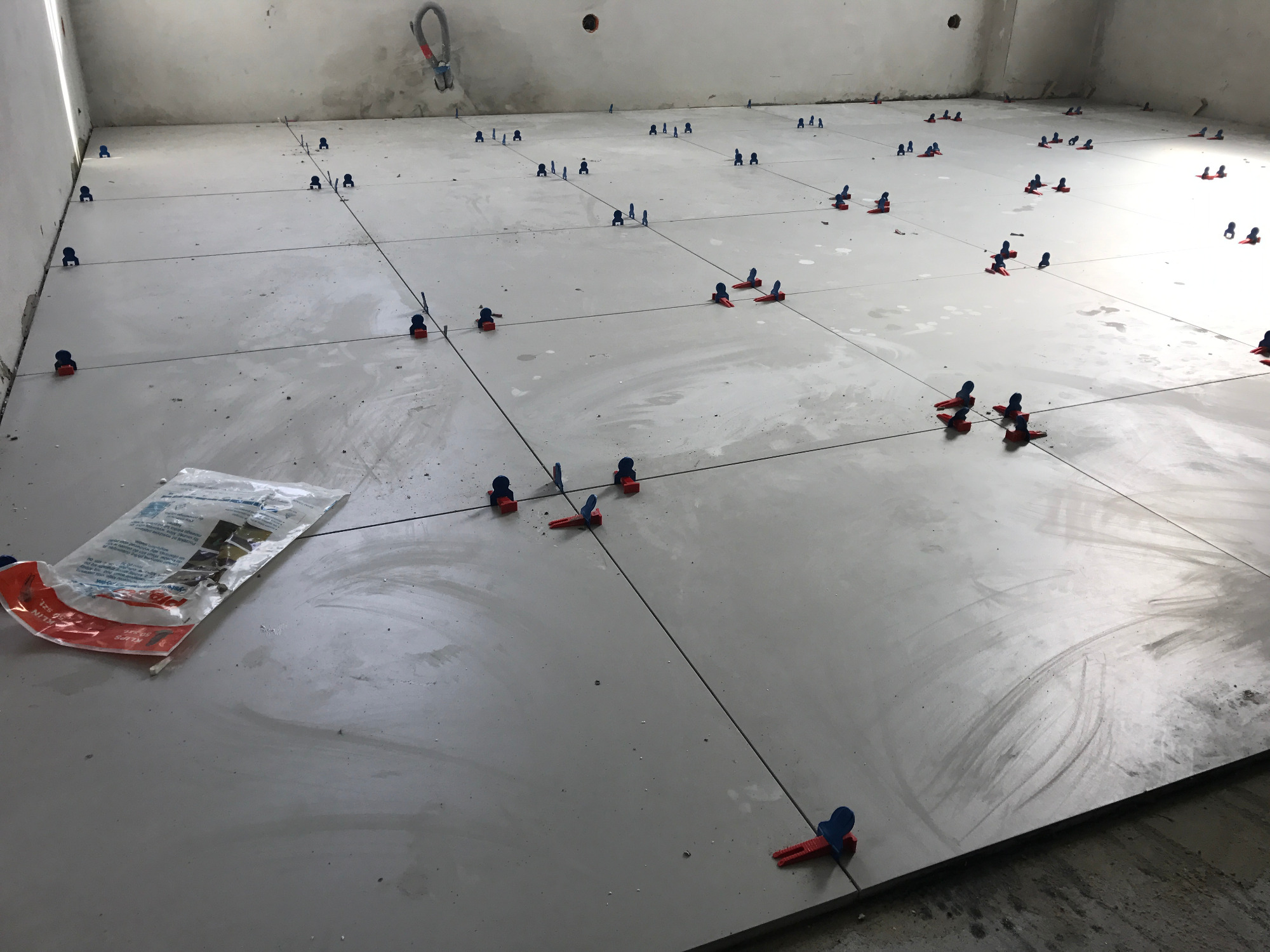 Układanie szarych płytek podłogowych z użyciem systemu poziomowania, widoczny worek z klinami i klamrami. W tle surowa ściana w trakcie remontu.