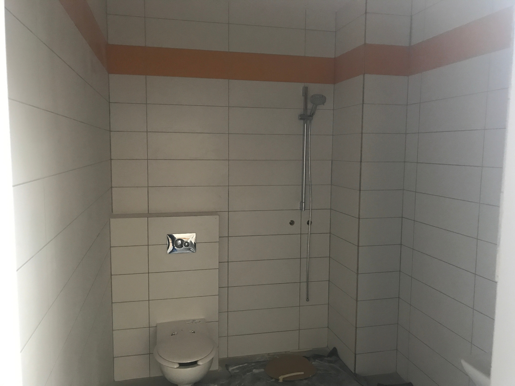 Wnętrze łazienki w trakcie remontu, widoczna toaleta, prysznic i białe płytki z pomarańczowym pasem dekoracyjnym, podłoga zabezpieczona folią.