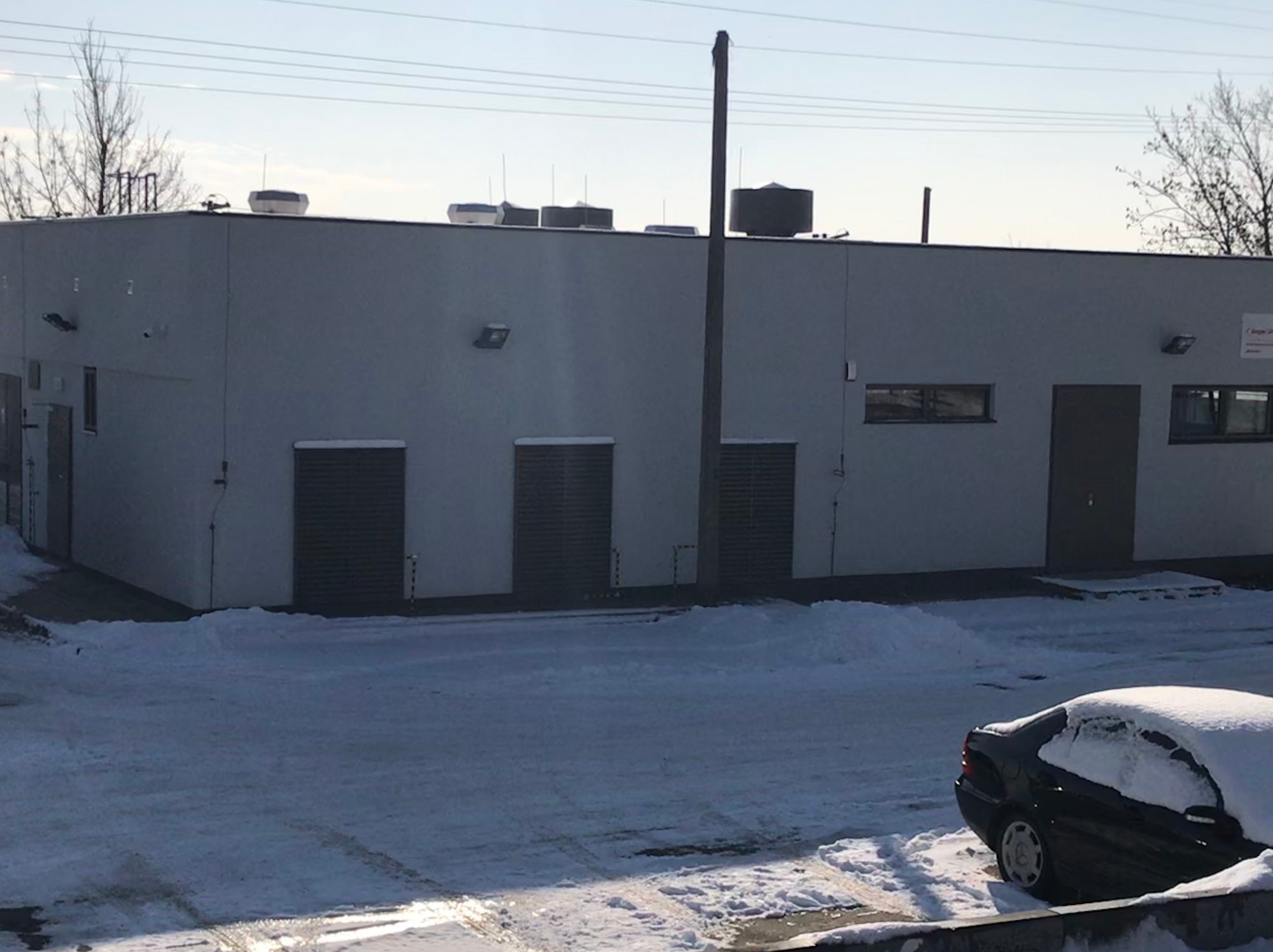 Szary budynek stacji podnoszenia ciśnień z kratami wentylacyjnymi, klimatyzacją na dachu i zaparkowanym, zasypanym śniegiem samochodem w zimowej scenerii.