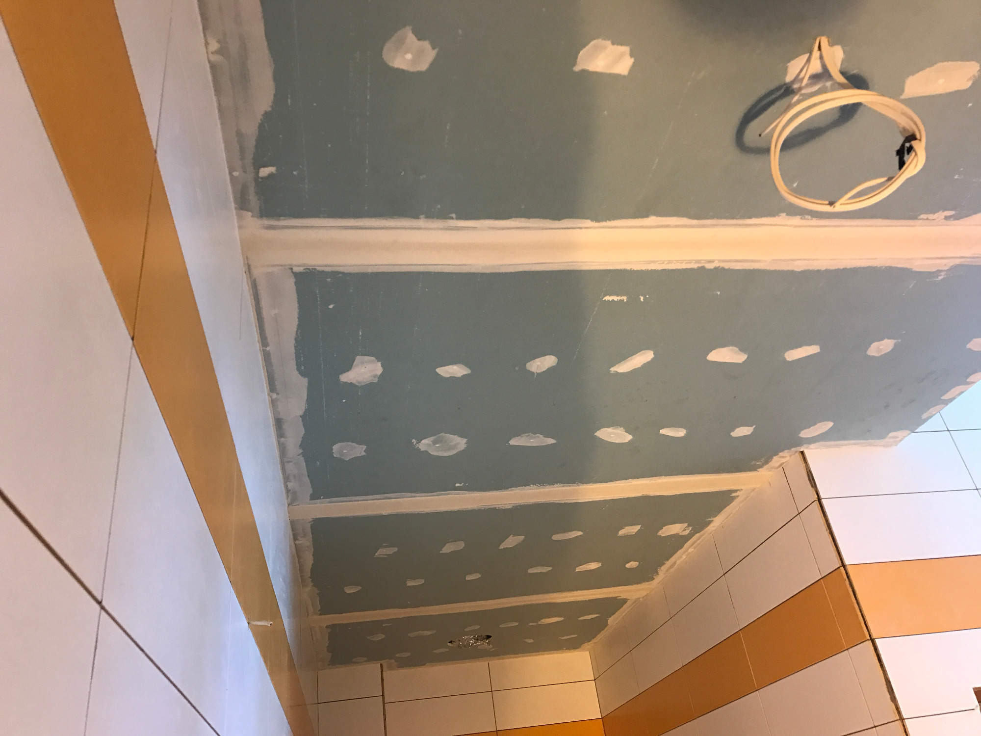 Niedokończony sufit podwieszany z płyt gipsowo-kartonowych w łazience z częściowo położonymi białymi i pomarańczowymi płytkami, widoczne kable elektryczne i szpachlowane łączenia płyt.