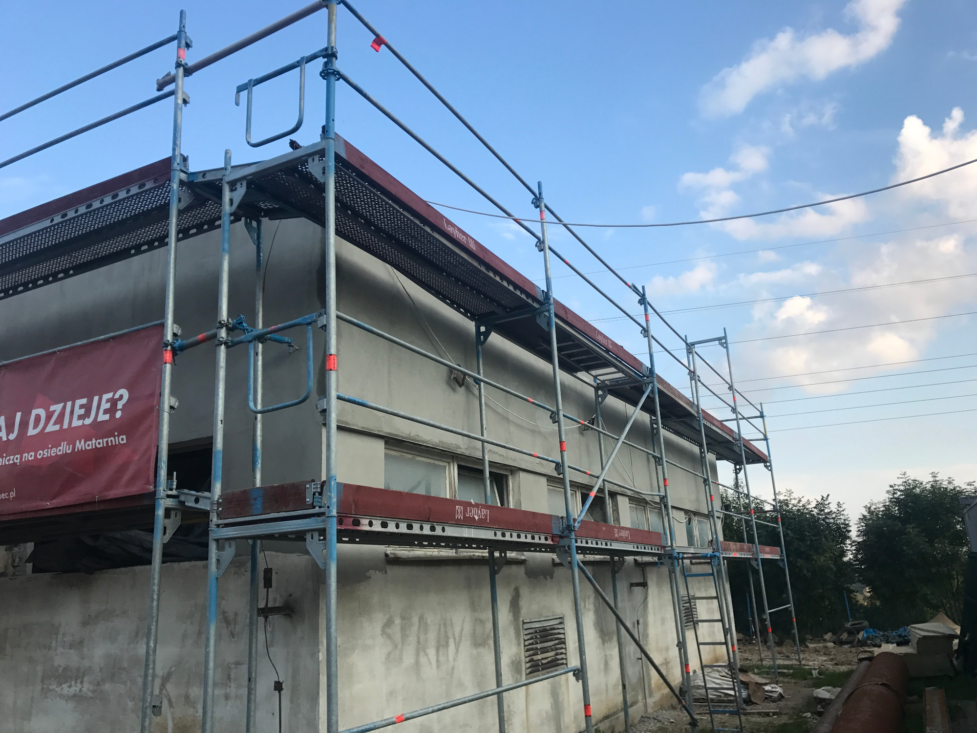 Budynek w trakcie renowacji, otoczony rusztowaniami Layher z czerwonymi akcentami, widoczny baner z napisem, błękitne niebo z chmurami w tle.