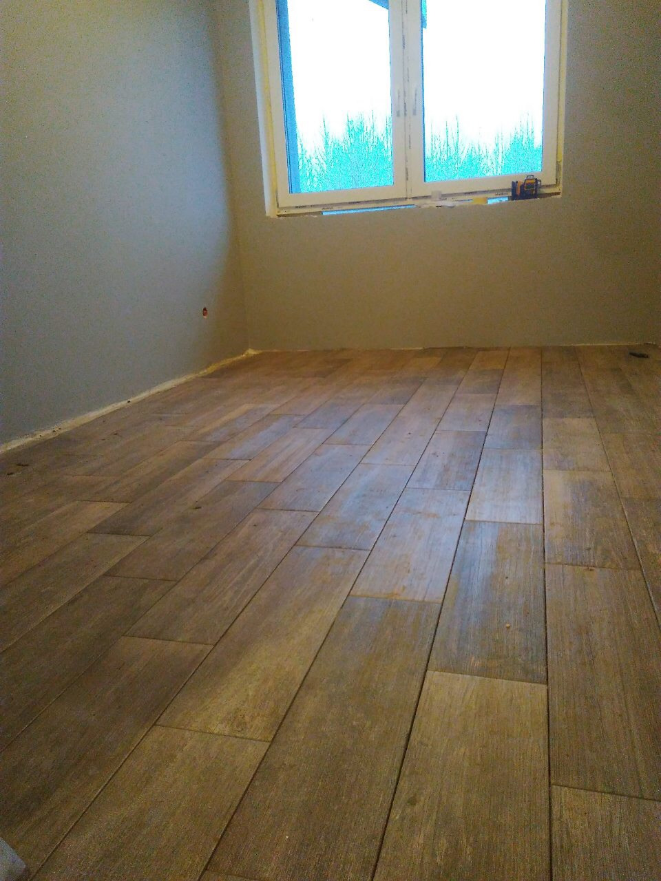 Ułożona podłoga z płytek imitujących drewno w nowym pokoju z szarą ścianą i oknem, widoczny poziomica na parapecie.