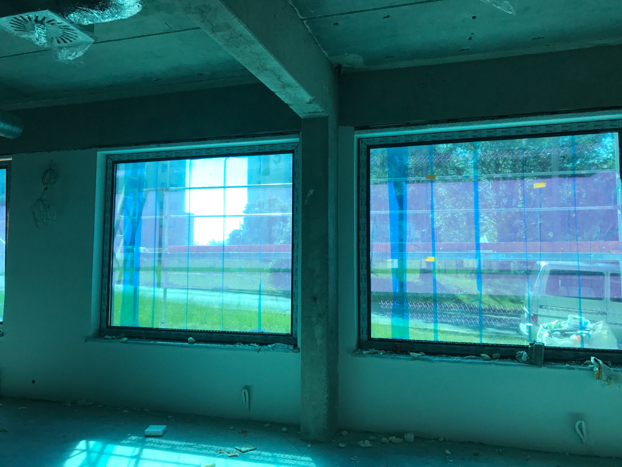 Wnętrze w trakcie remontu z dwoma oknami zabezpieczonymi folią, widoczne surowe ściany i betonowy filar, pozostałości pianki montażowej przy oknach.