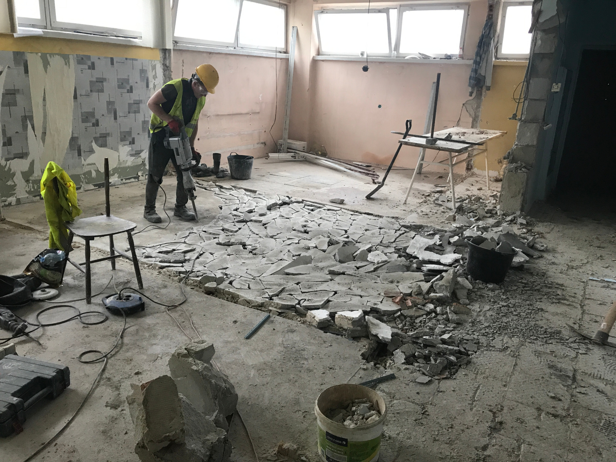 Pracownik w kasku i kamizelce odblaskowej używa młota pneumatycznego do skuwania posadzki w pomieszczeniu w trakcie remontu. Widoczne gruz, narzędzia i przedmioty zabezpieczające.