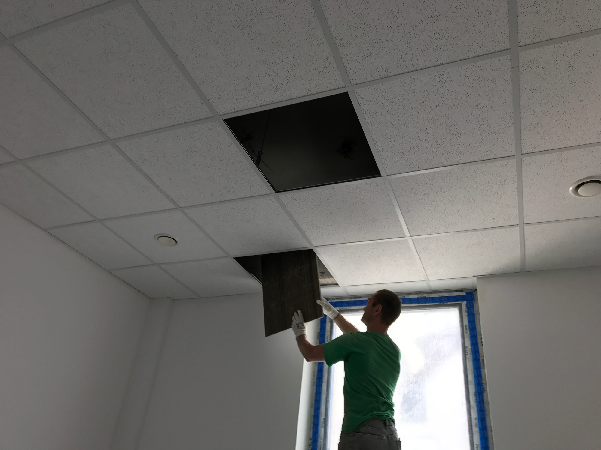 Pracownik w Nowym Dworze Gdańskim montuje kaseton sufitowy w pomieszczeniu z oknem, widoczny fragment ściany i biała kratownica sufitu podwieszanego.