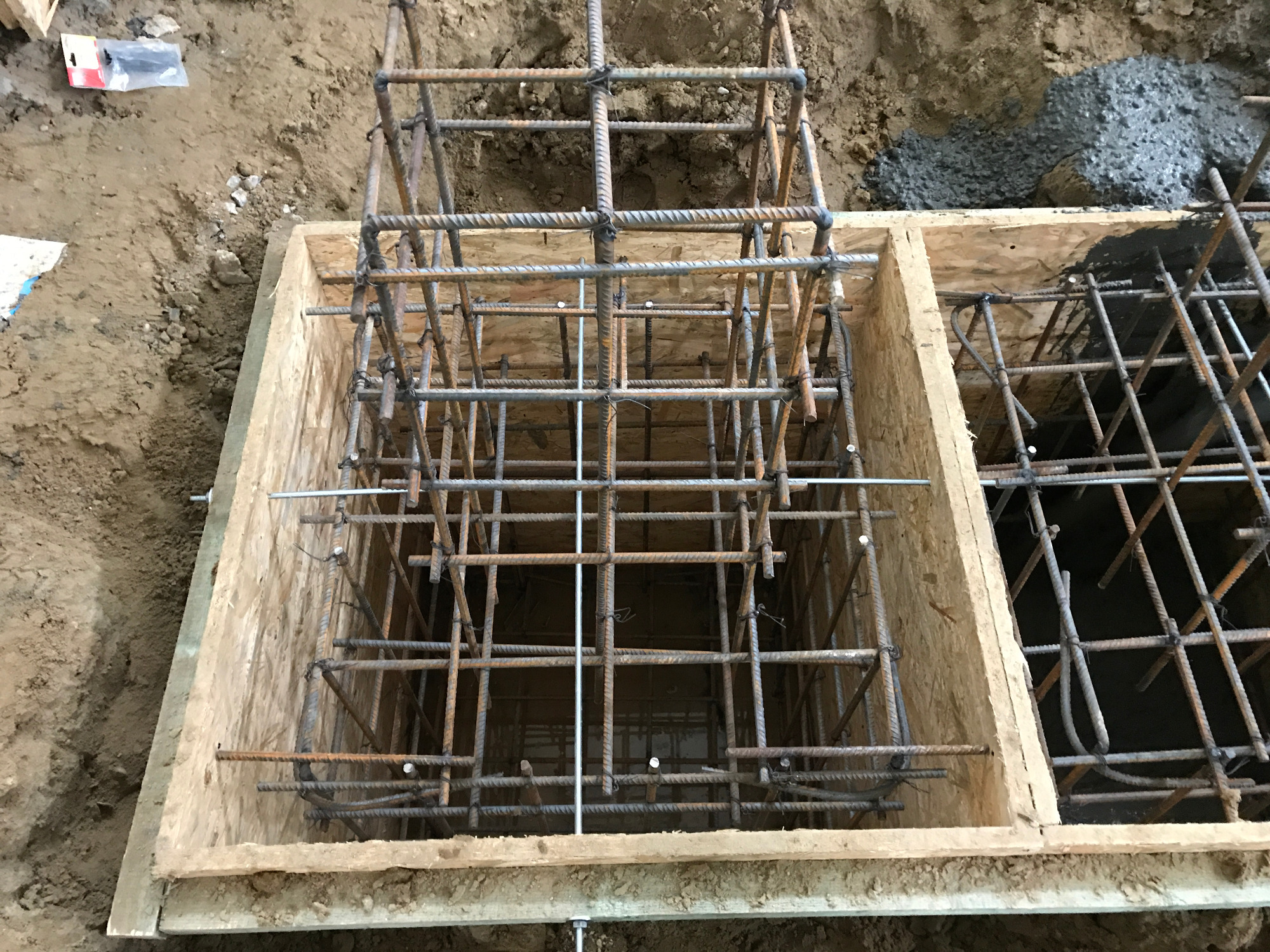 Zbrojenie fundamentu w drewnianym szalunku, widoczne pręty zbrojeniowe połączone drutem, przygotowane do zalania betonem. Częściowo zalany beton w sąsiedniej sekcji szalunku.