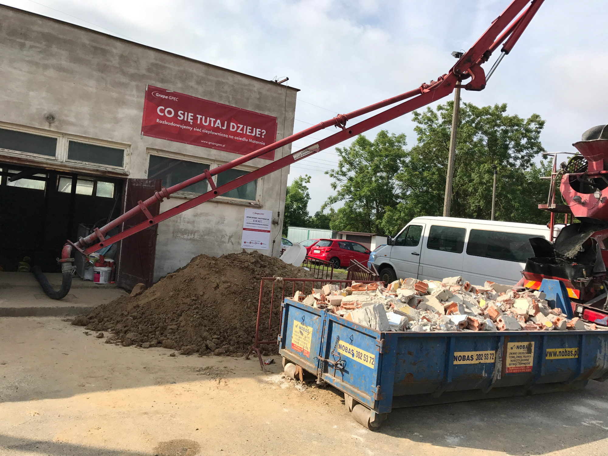 Renowacja stacji podnoszenia ciśnień w Gdańsku: widok na budynek z czerwonym banerem Grupa GPEC, wykop ziemi, kontener z gruzem oraz ramię pompy do betonu.