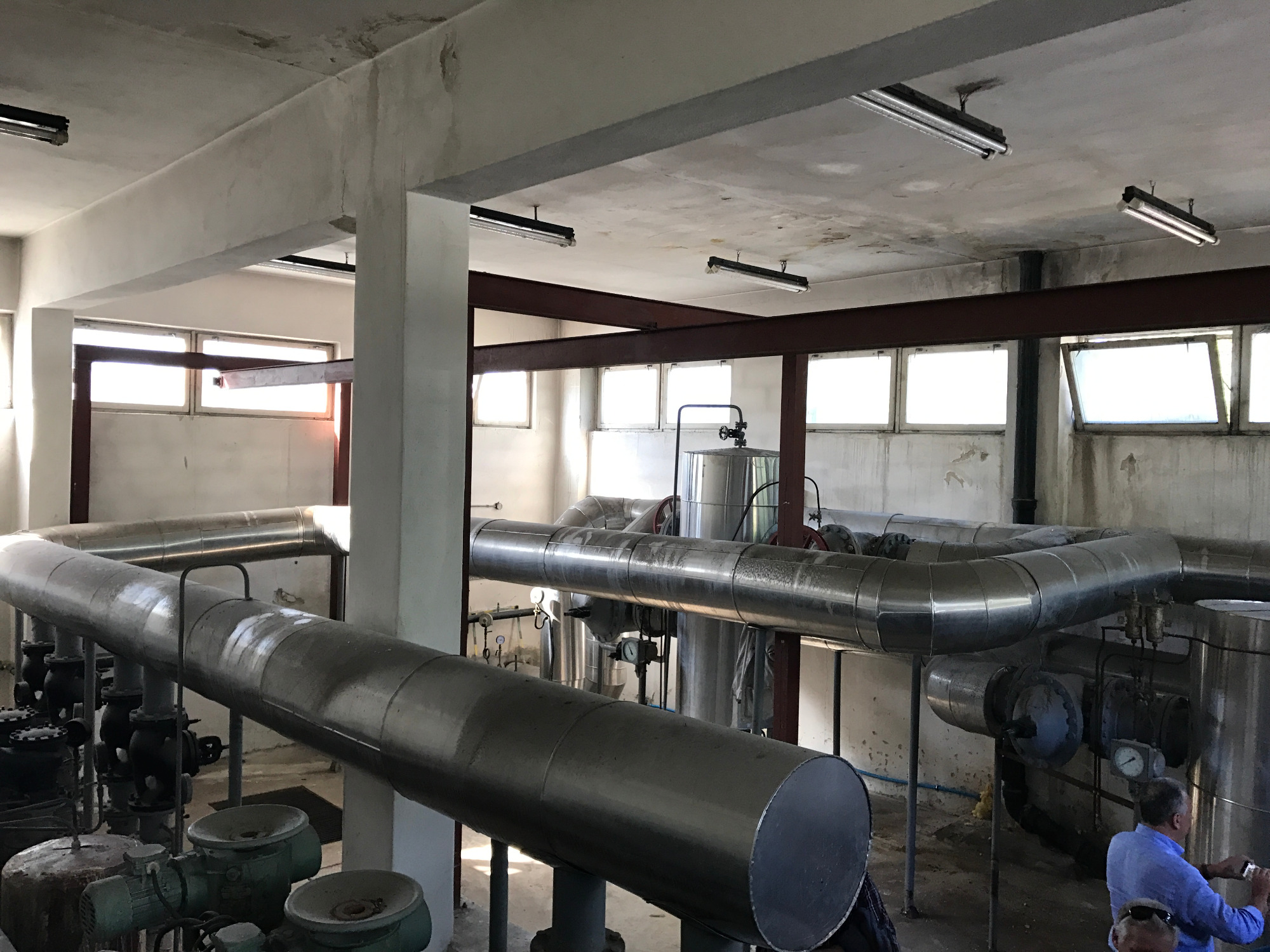 Wnętrze stacji podnoszenia ciśnień z widocznymi rurami, zbiornikami i elementami instalacji, oświetlone przez okna i lampy sufitowe, z fragmentem osoby w prawym dolnym rogu.