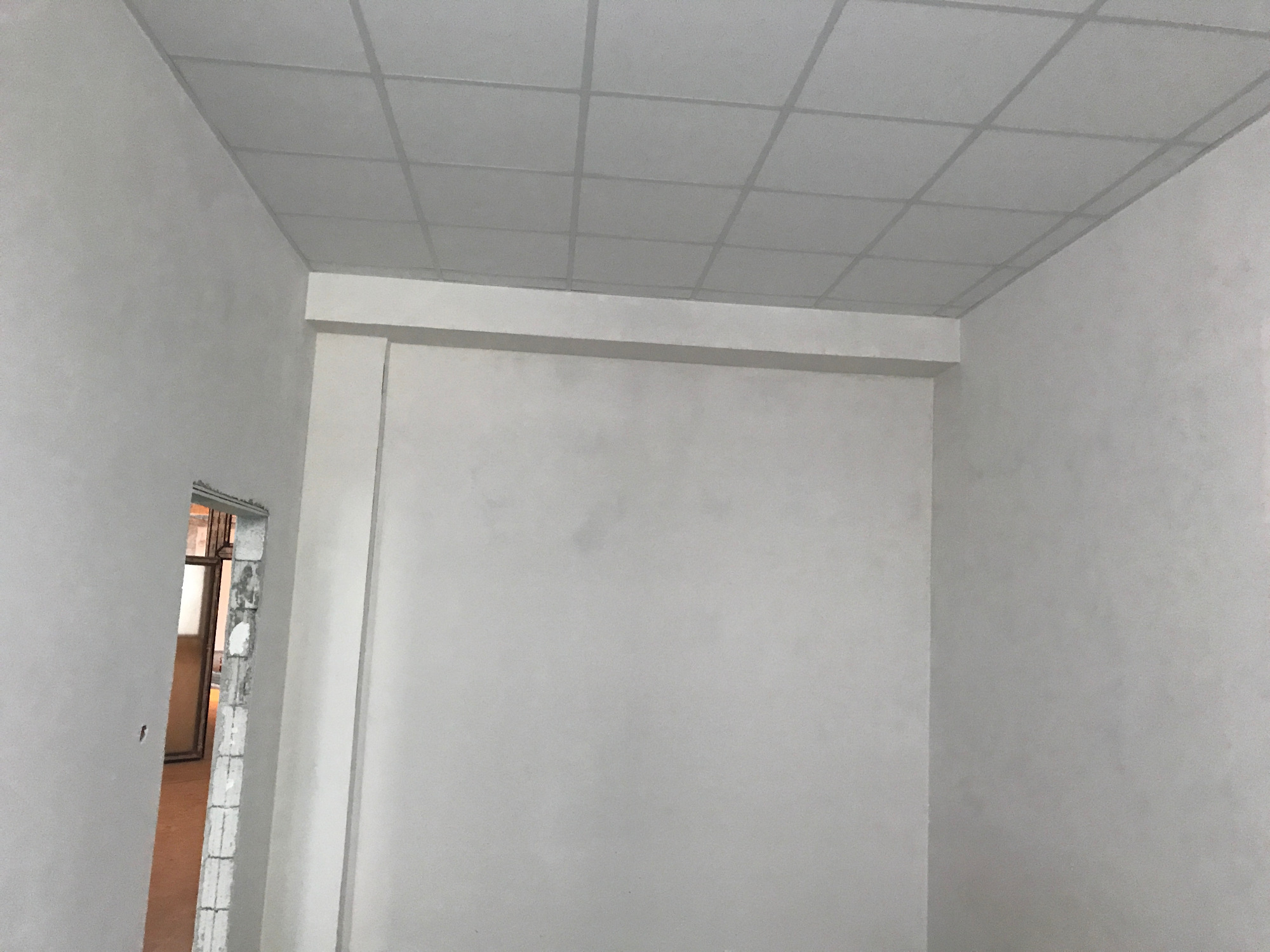 Wnętrze w trakcie remontu: białe ściany, sufit podwieszany z kratownicą, widoczne ślady po pracach budowlanych przy ościeżnicy drzwiowej.