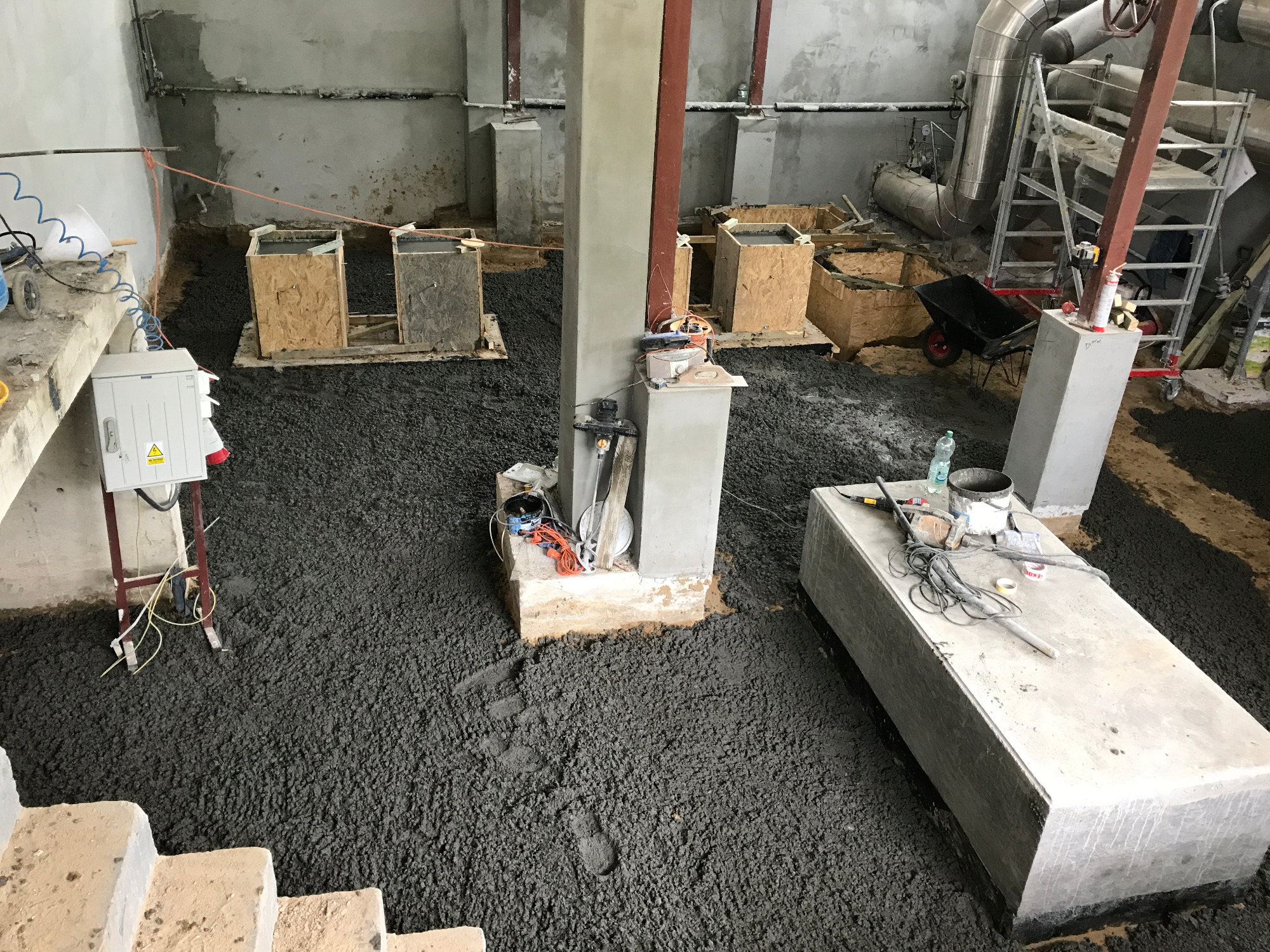 Wnętrze budynku w trakcie wylewania posadzki z ciemnego betonu, widoczne formy szalunkowe, narzędzia, drabina i instalacje pod sufitem.