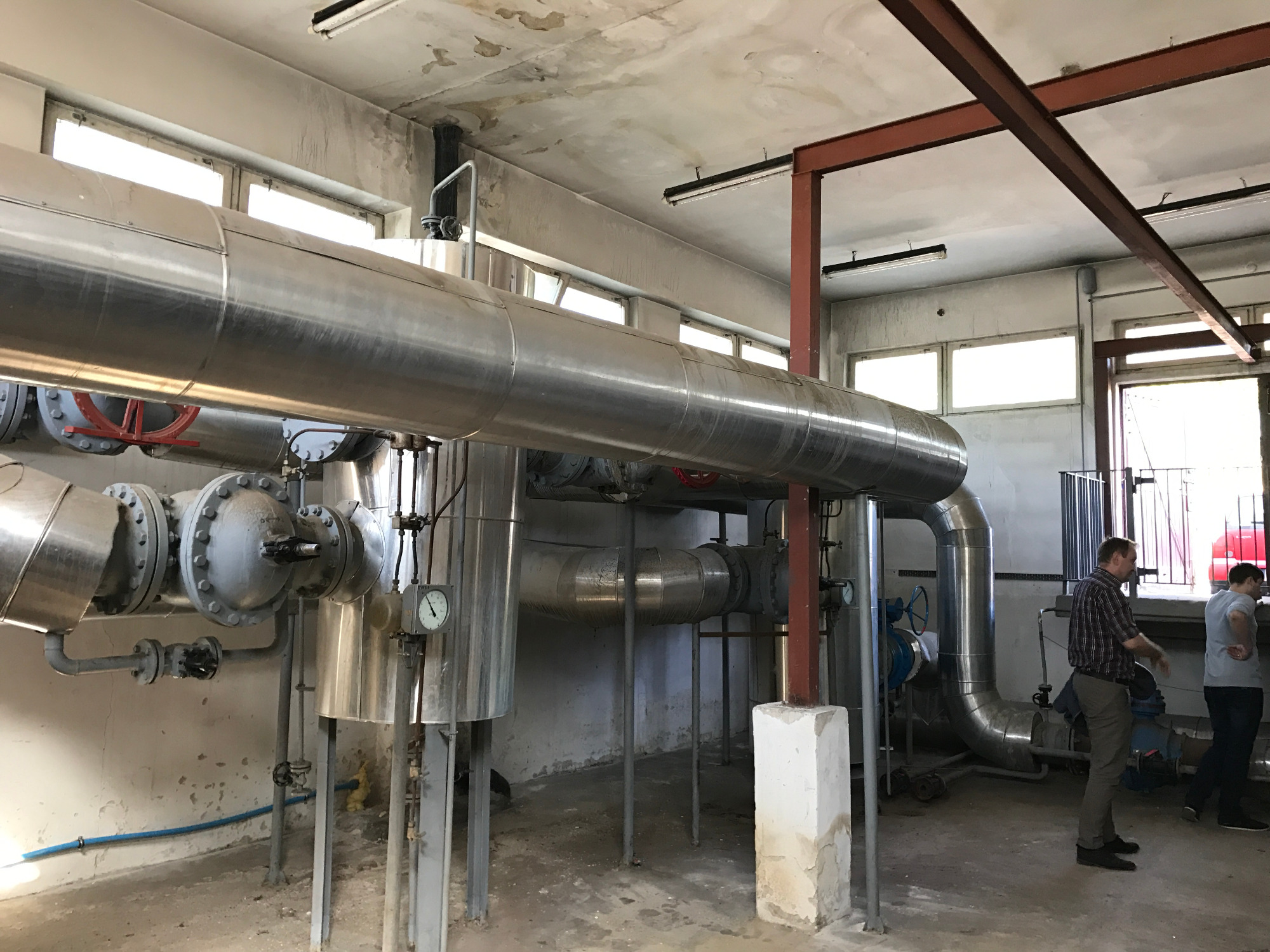 Wnętrze stacji podnoszenia ciśnień w trakcie renowacji: widoczne rury ze stali nierdzewnej, manometry, fragmenty instalacji, dwóch mężczyzn w tle, surowy betonowy sufit z widocznymi śladami wilgoci.