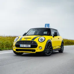 Ż&oacute;łty Mini Cooper z czarnymi felgami i oznaczeniem kursanta nauki jazdy na dachu, zaparkowany na szosie z polem kukurydzy w tle.