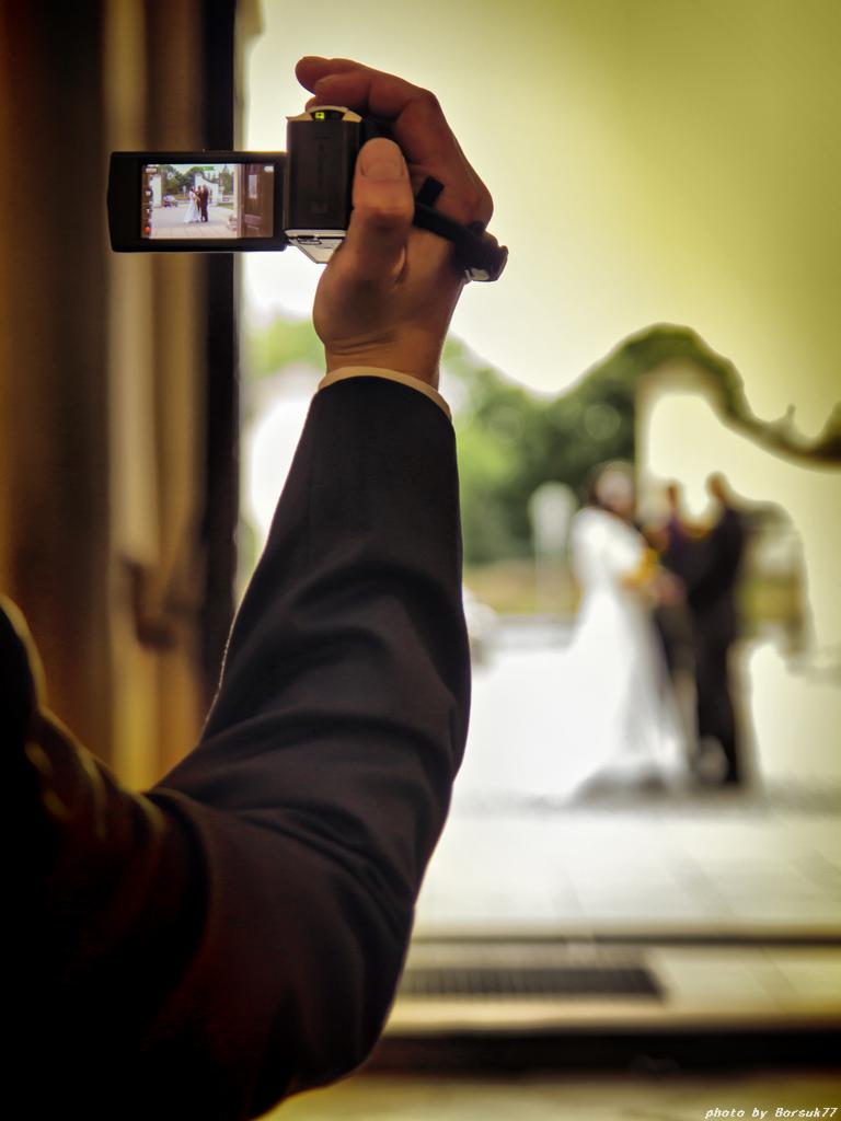 Ręka trzyma kamerę wideo z widokiem na parę młodą podczas ceremonii ślubnej, zamazane tło z zielenią i dekoracją.