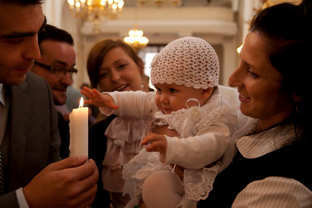 Uśmiechnięte niemowlę w białym, koronkowym ubranku i czapeczce, trzymane przez kobietę, wyciąga rączkę w kierunku płonącej świecy trzymanej przez mężczyznę w garniturze, w tle widoczni inni goście...