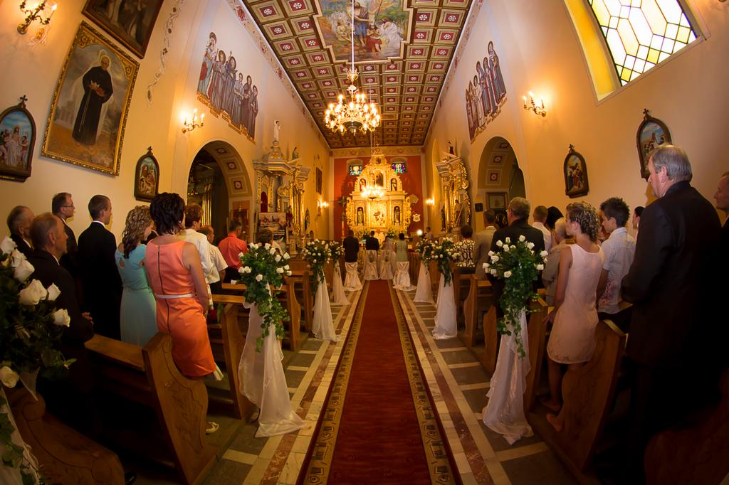 Wnętrze kościoła udekorowane białymi kwiatami i tiulem wzdłuż ławek, przygotowane na ceremonię ślubną, widziane z perspektywy gości.