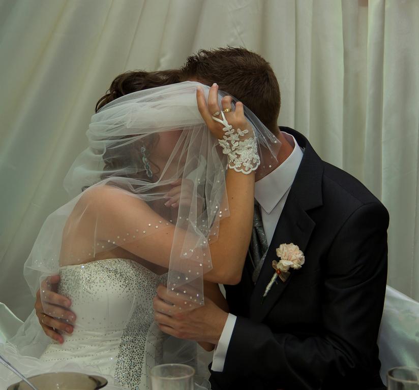 Panna młoda w sukni z błyszczącym gorsetem i koronkowych rękawiczkach, całowana przez pana młodego w ciemnym garniturze z butonierką, zasłonięta welonem.