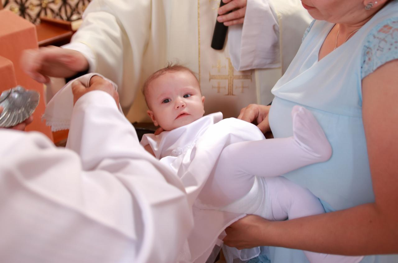 Ujęcie z uroczystości chrztu dziecka, z widocznym księdzem polewającym wodą główkę niemowlęcia, trzymanego przez matkę w jasnoniebieskiej sukience.